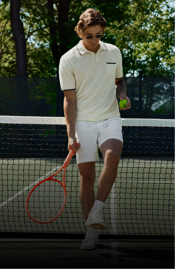 Man on a tennis court holding a racket and ball, wearing a white polo shirt and shorts.