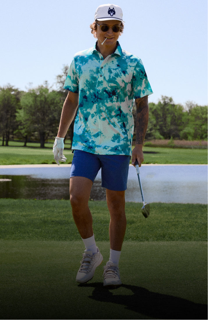 Man in a tie-dye shirt and blue shorts walking on a golf course.